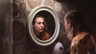 En bild från HBO Max-serien Welcome to Utmark, där en kvinna kollar sig i en rund spegel i ett mörkt badrum.