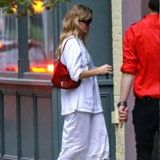 Jennifer Lawrence enters into a building wearing a white lace-trim slip skirt with a white baggy t-shirt and white strappy sandals. She wears a red suede bag over her shoulder.
