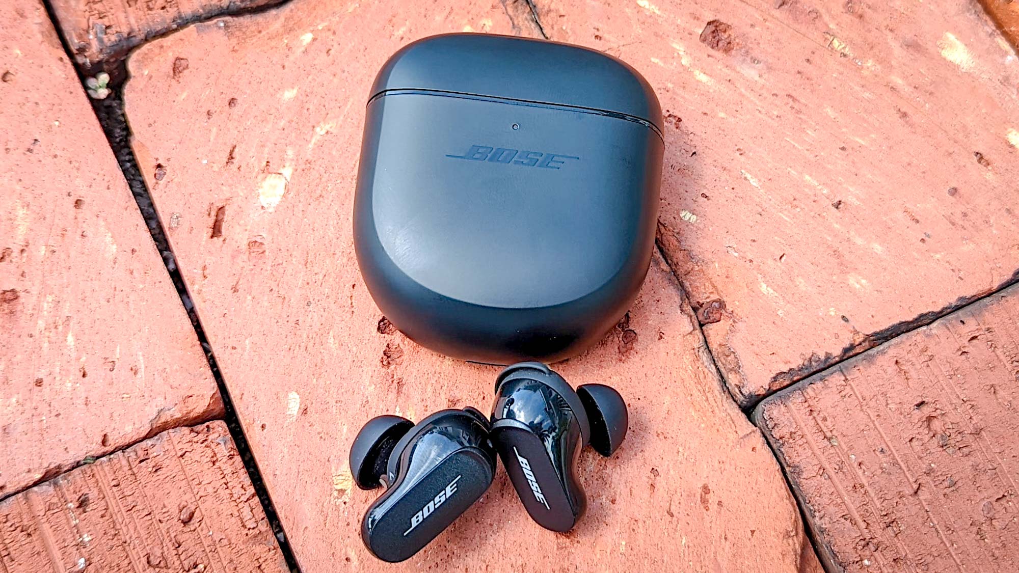 Bose QuietComfort Earbuds 2 en color negro con estuche de carga colocado sobre pavimento de ladrillo