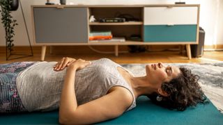 Woman lying on her lack on a yoga mat on the floor of the living room, eyes closed, meditating and practicing breathwork as a way to get back to sleep