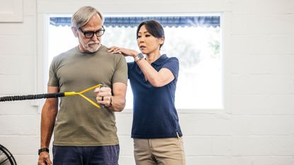 A trainer stands behind a client assisting him with an exercise. The trainer is touching the client's right shoulder, while the client holds the end of a resistance band in his right hand, with his elbow bent at 90 degrees and pinned to his side.