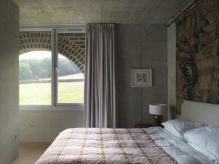 A bedroom with a bed, red checkered linen, a side table, a lamp, a wall tapestry and a large window.