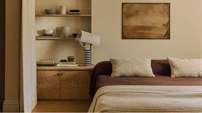 cream bedroom with artwork above bed, brown sheets with cream cushions and throw, a textured dimpled timber side table with floating shelves styled with vases above, a striped table lamp, a curtain