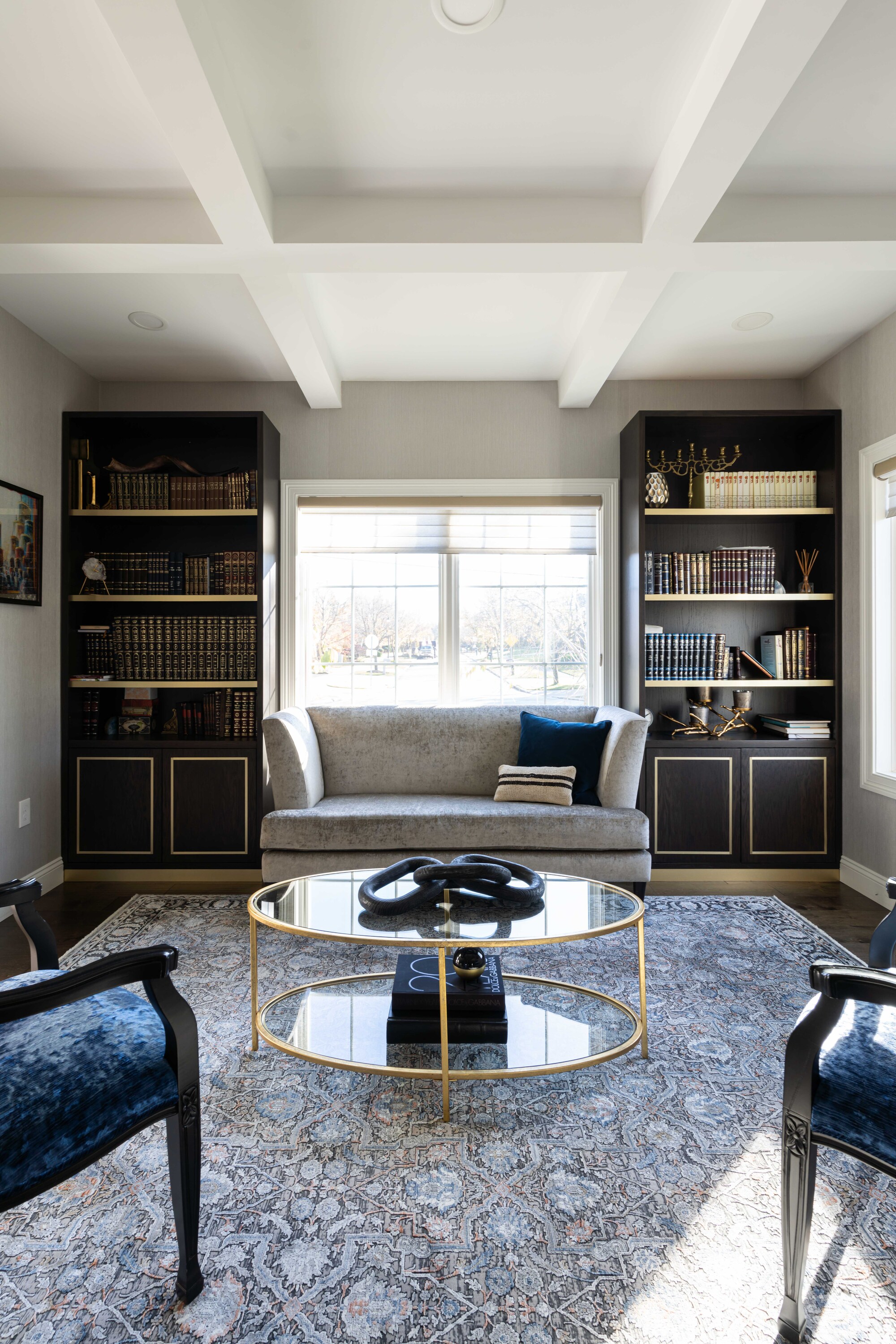 A large living room with two tall bookcases flanking a window, a round glass coffee table, and a large rug
