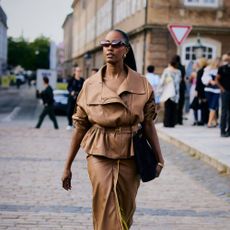 Woman wears cropped tan trench and matching skirt with a handbag as she walks through the street. 