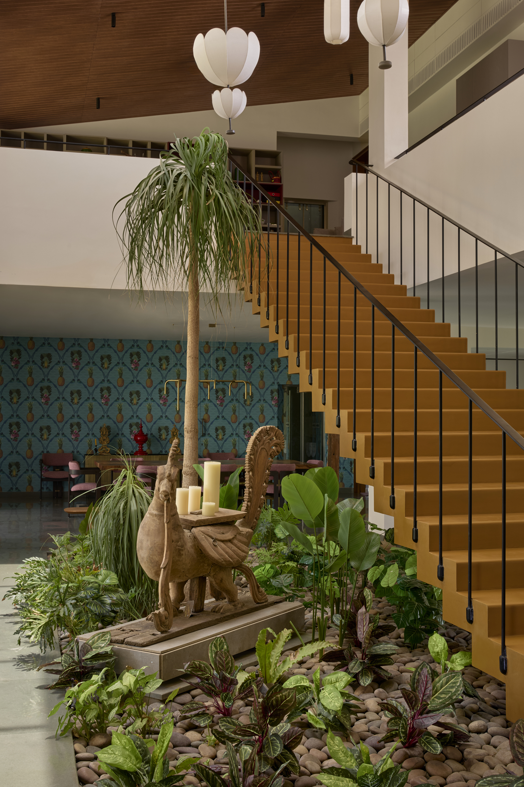timber staircase in the lobby of home with a lush indoor garden at its base with plants and stones and a tree. upstairs there is a library and white pendant lights, and downstairs there is a blue patterned wallpaper room and polished concrete floors