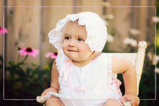 A beautiful baby girl, sixteen months old, sits in an antique chair, wearing a vintage dress and bonnet