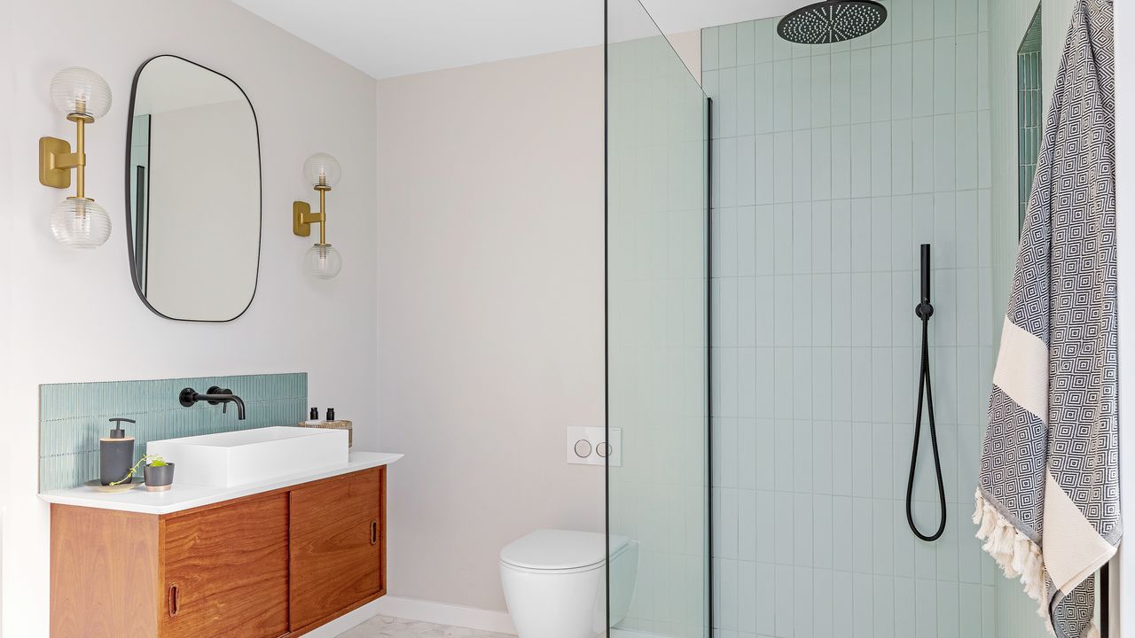 A modern bathroom with matt black showerhead and fitxture, teal green wall tiles, marble hexagonal floor tiles, teak wood vanity, rectangular ceramic bathroom basin, soft rectangular mirror, and pair of brass light fixtures on white walls