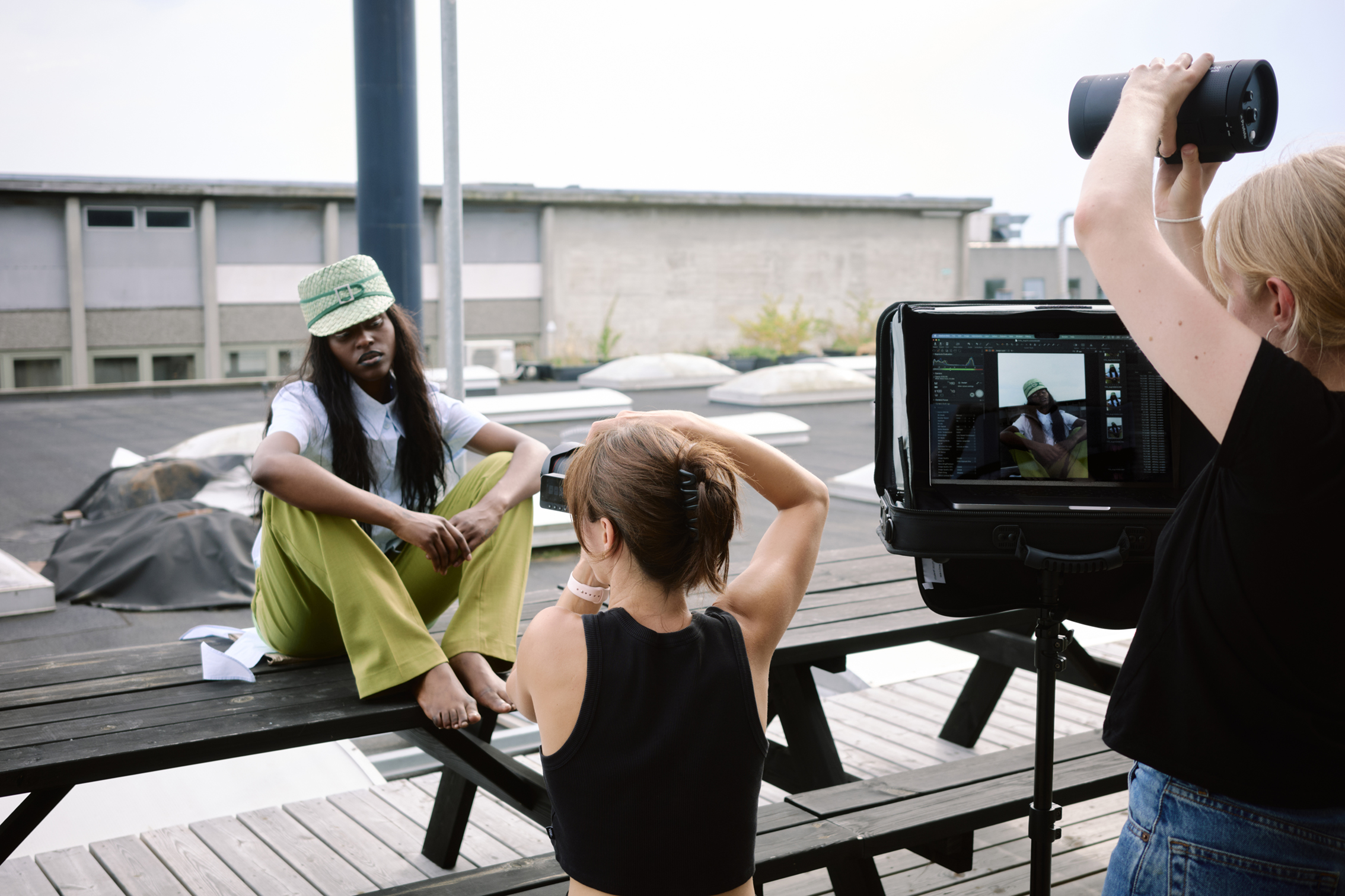 Photo shoot on rooftop with barefoot model in green pants and matching hat sitting on bench while a photographer captures her image
