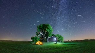 A man with a lantern next to his camping tent is watching the Perseids meteor shower under a great century-old pine tree. Starry sky panorama with The Perseid meteor shower in a agricultural field. Milky Way on the background