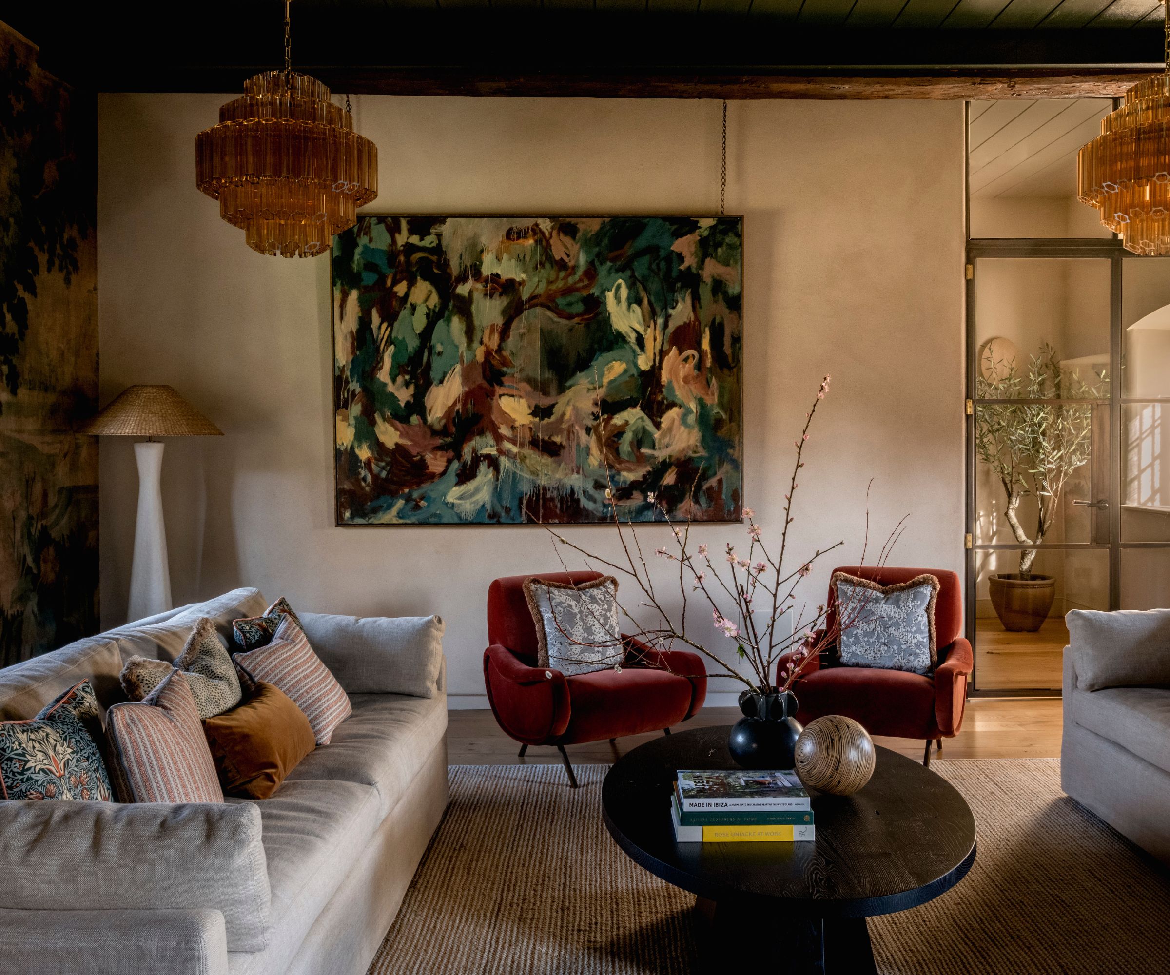 earthy moody living room with neutral walls, a large green abstract painting, two neutral couches facing each other, a dark wooden round coffee table and two rust red velvet chairs