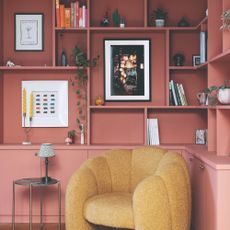 A built-in bookcase with bottom doors painted in a terracotta shade with a yellow boucle accent chair in front of it