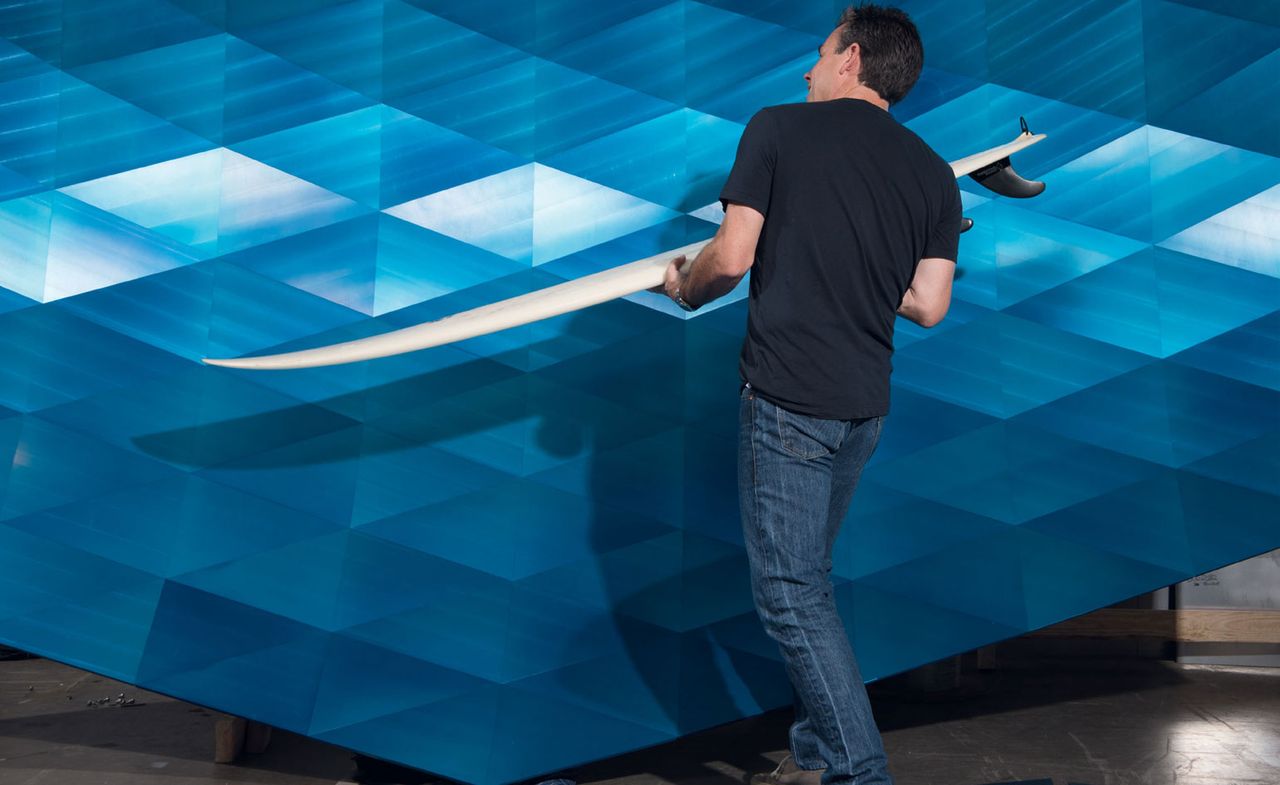 View of Alex Rasmussen standing in front of &#039;The Wave&#039; installation which features blue anodised aluminium triangles. He is wearing a dark blue t-shirt and jeans and he is holding a white surfboard in his hands