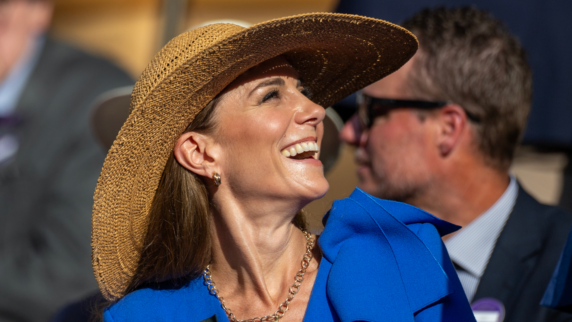 Kate Middleton wearing a blue dress and a straw hat looking up and laughing