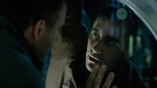 This is a still from the movie Life (2017). Here we see a close up of two men looking intensely at each other through a window. One man has his hand up and placed on the window.