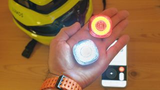 A user's hand shows two Lumos Firefly lights with the companion app and Lumos Ultra Fly Pro helmet out of focus in the background