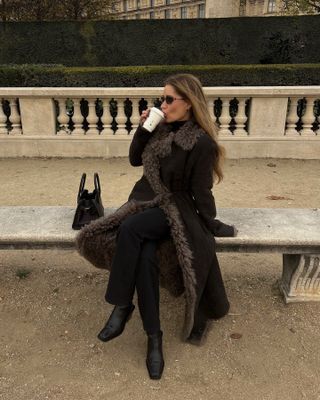 A woman wearing a black fur coat, black straight-leg jeans, black ankle boots, and retro-inspired sunglasses.