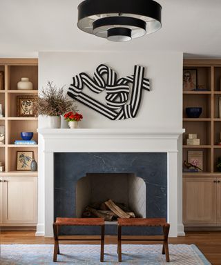 Mantelpiece with vase of foliage, vase of flowers and artwork above, two stools in front on area rug