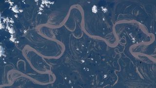 An image of the Ucayali River taken from space shows the river snaking in a light brown color across dark blue and green land with some small fluffy white clouds overhead. 