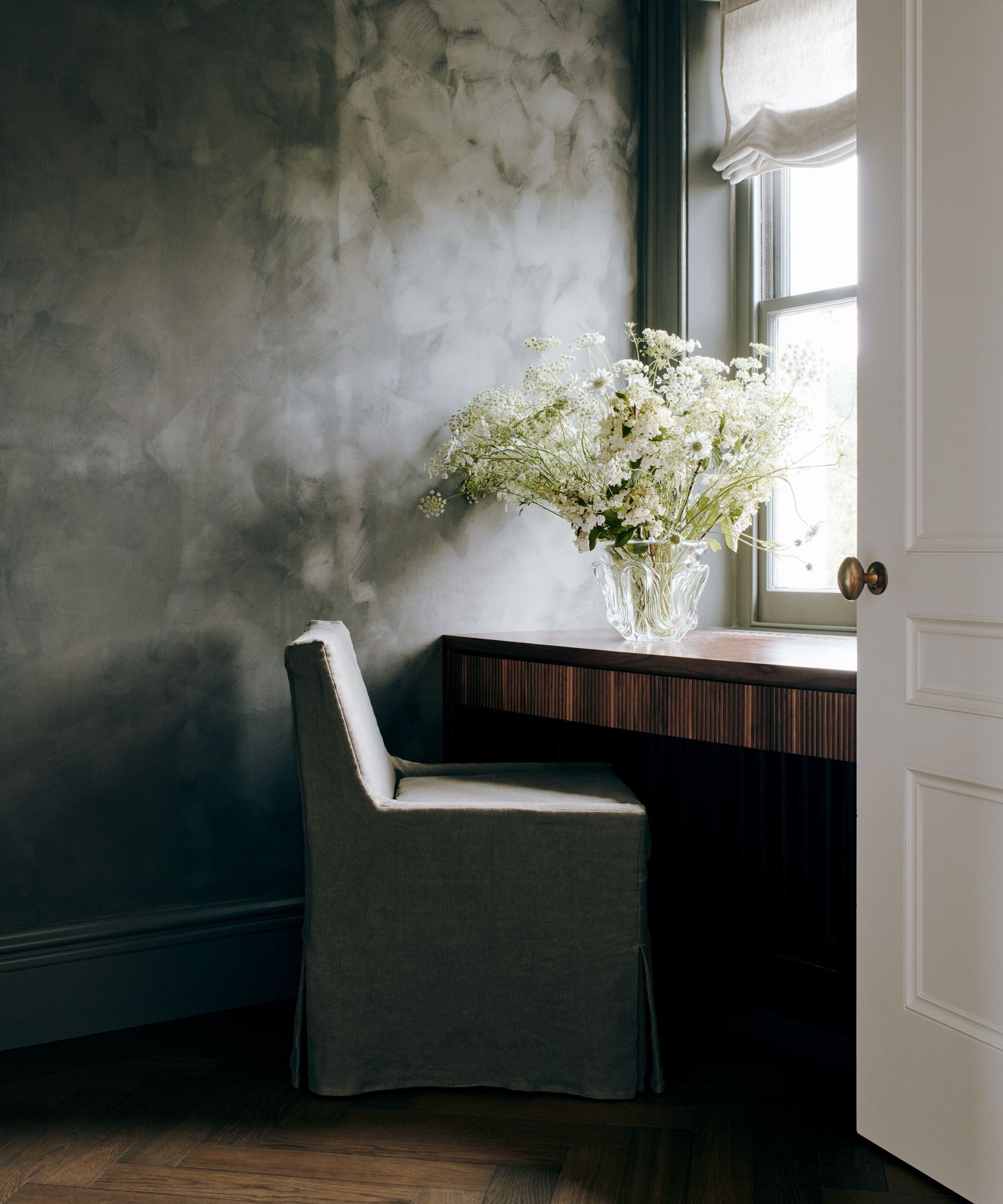Wooden desk in front of windiw, vase of flowers on top, gray textured walls, dark wood floor