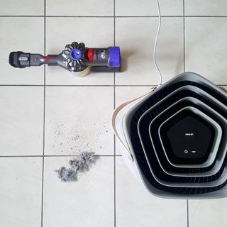 The AEG AX91-604GY Connected Air Purifier on a tiled floor with a pile of dust and a Dyson vacuum cleaner next to it