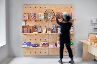 A silver room with a wooden desk, wooden wall shelving with books on them and a person standing in front of it.