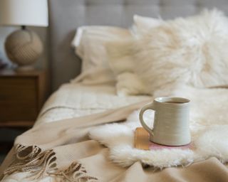 a cozy bed closeup with fuzzy pillows and a throw blanket and a mug at the foot of the bed