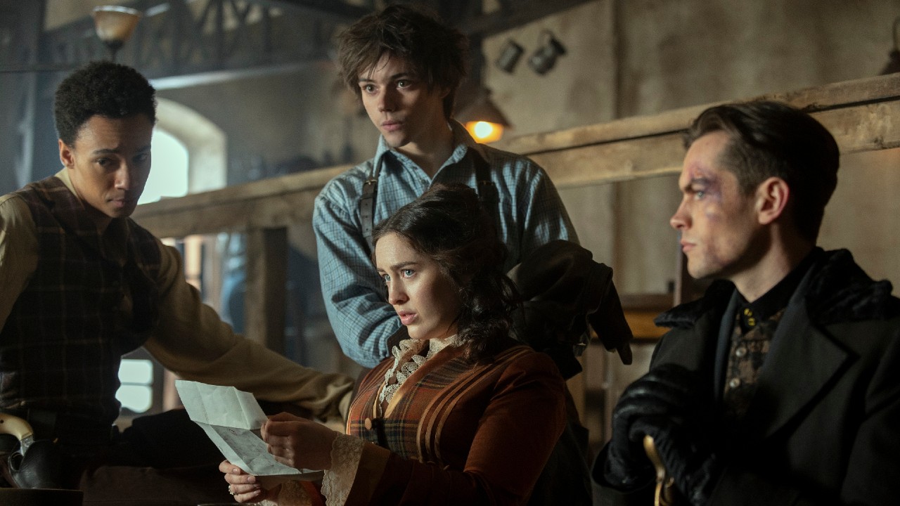(L to R) Kit Young as Jesper Fahey sitting on a table, Danielle Galligan as Nina Zenik holding a piece of paper with both hands and looking up, Jack Wolfe as Wylan standing behind her, Freddy Carter as Kaz Brekker sitting next to her.