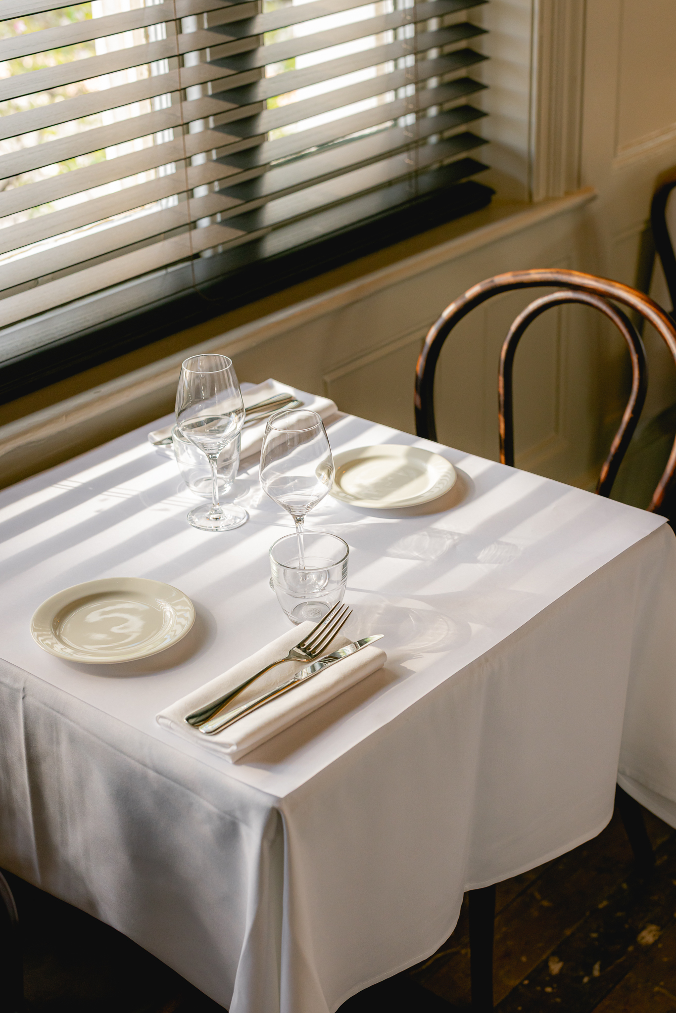 The charming-cool interiors of a French-style modern bistro with distressed plaster walls, lived-in wooden floors, elegant tables and chairs, colorful artworks, beige and red curtains, and sunset-hued sconces and pendant lights.