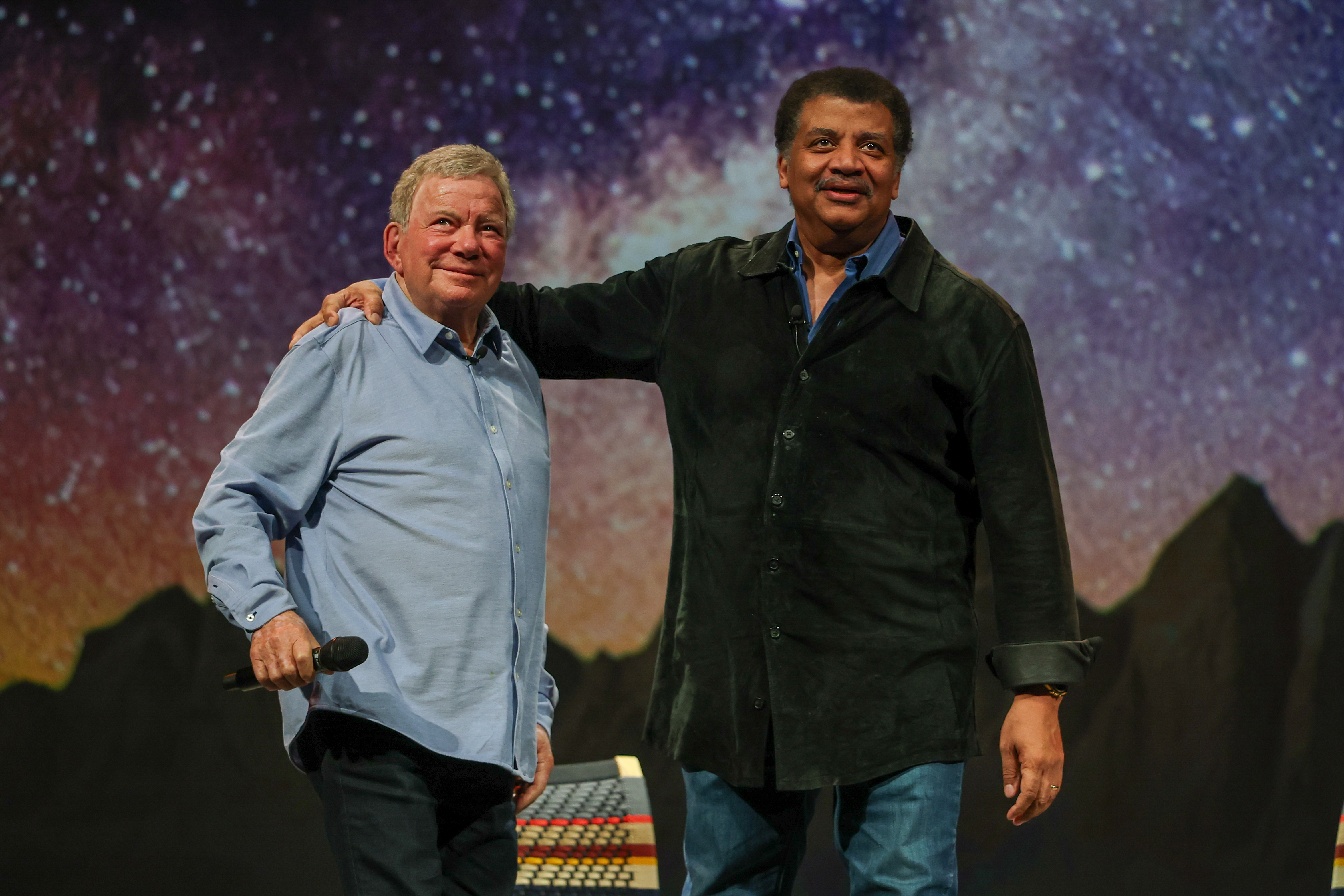 A man wearing a blue shirt stands to the left of a man wearing a black shirt, both in front of a starry background