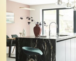 black marble kitchen island, pink and yellow walls, terracotta vase, emerald green chairs, glass pendant lights