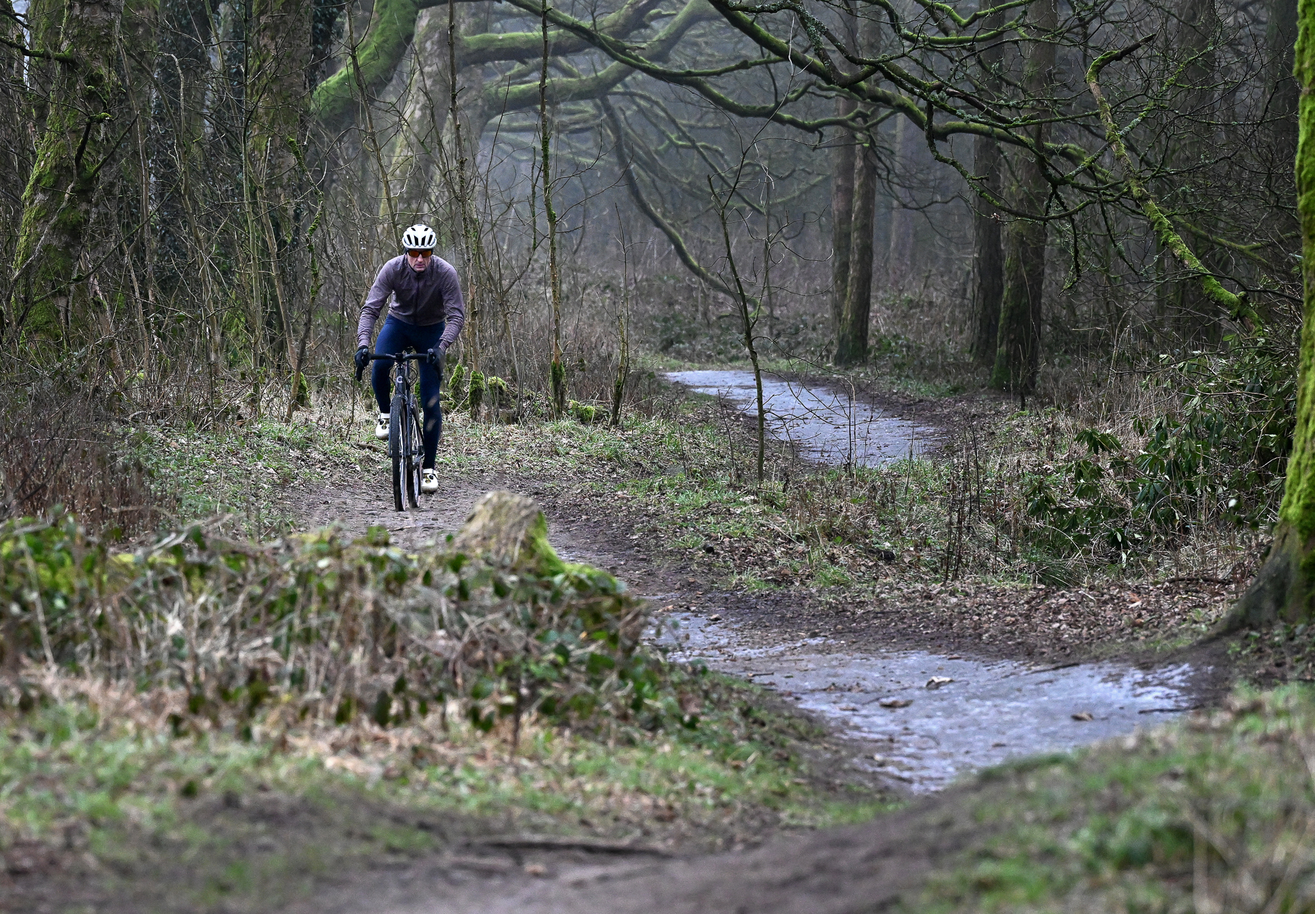 front view of a man wearing navy tights and a purple jacket riding a black gravel bike on a dirt trail towards the camera on a wintry day