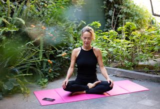 A woman meditating on top of a pink yoga mat while sat outside in her green, leafy garden.