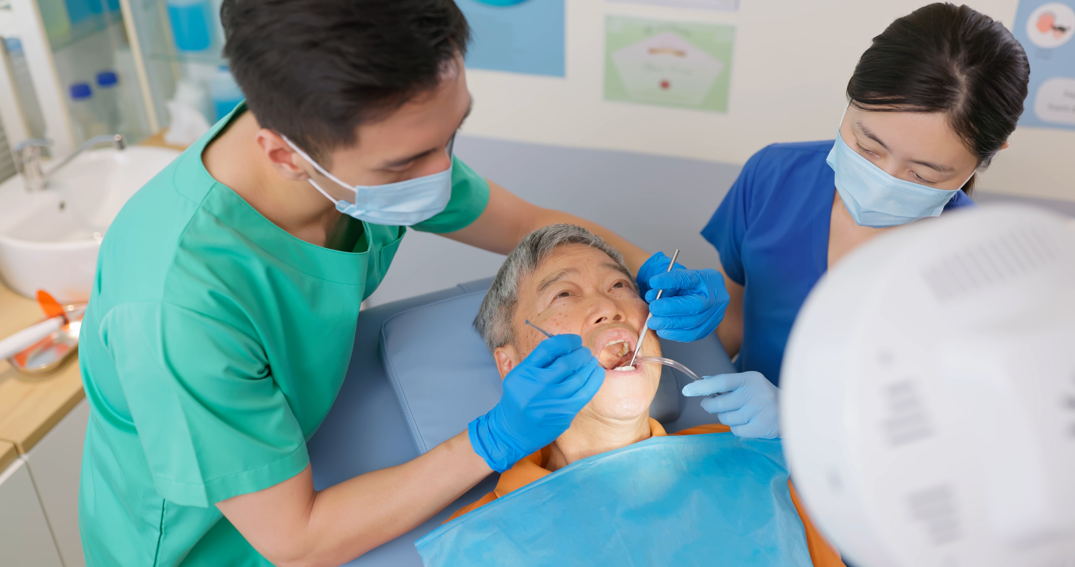 top view asian male dentist examining a elderly man patient teeth as he lies on dental chair in the clinic with nurse assistance aside