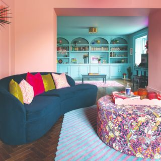 A colourful living room painted in pink with a curved blue sofa, a striped and scalloped light blue rug and a large round floral ottoman
