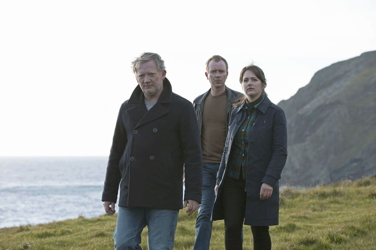 A group shot of the three lead investigators on Shetland: DI Jimmy Perez (Douglas Henshall), DC Sandy Wilson (Steven Robertson) and DS Alison &#039;Tosh&#039; Mackintosh (Alison O&#039;Donnell). They are standing on a grassy clifftop with the sea in the background. Perez is wearing a black peacoat over a grey sweater and blue jeans, Sandy is wearing a black leather coat over a brown jumper and blue jeans, and Tosh is wearing an long open dark jacket over a green-and-black plaid shirt paired with a green rollneck, and black trousers