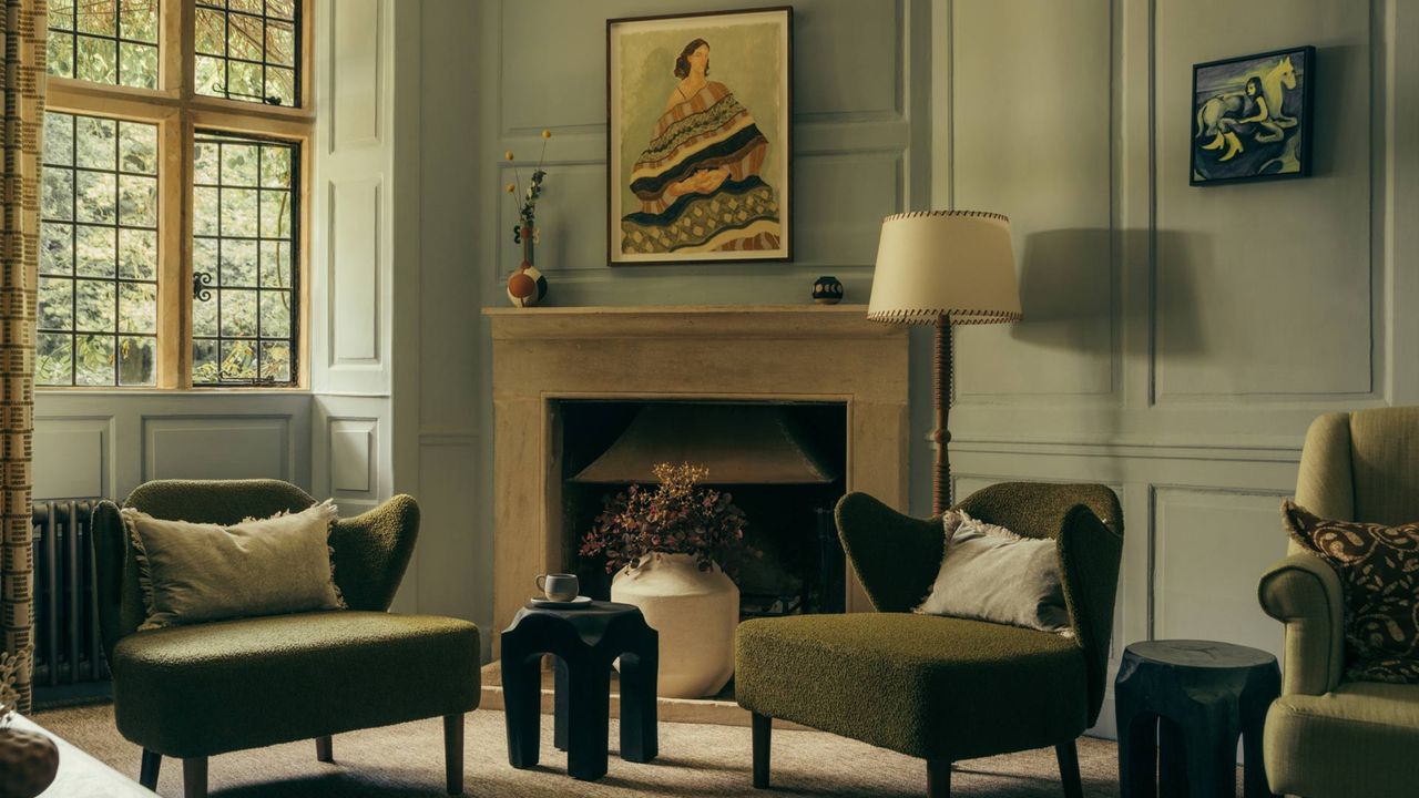 A vibrantly decorated farmhouse with pale blue wooden walls, upholstered mid-century furniture mixed with Liberty-style one in moss tones, figurative artworks depicting women, carpeted floors, and a warm ambiance.