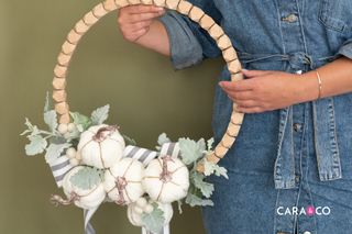 fall wreath DIY with wooden wreath ring, pumpkins, faux greenery held by a person
