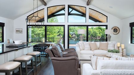 Open plan kitchen ideas are so chic. Here is a black and white breakfast bar with white stools, brown and white couches, and a large cut-out arched window with a view of trees and a river