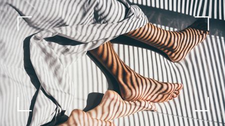 Bird's eye view of couples feet in bed during hot morning with sun coming through slatted blinds, representing how to sleep in the heat