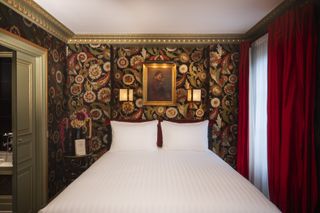 A gold-framed artwork on a wall covered in thick wallpaper, next to red curtains and a white bed