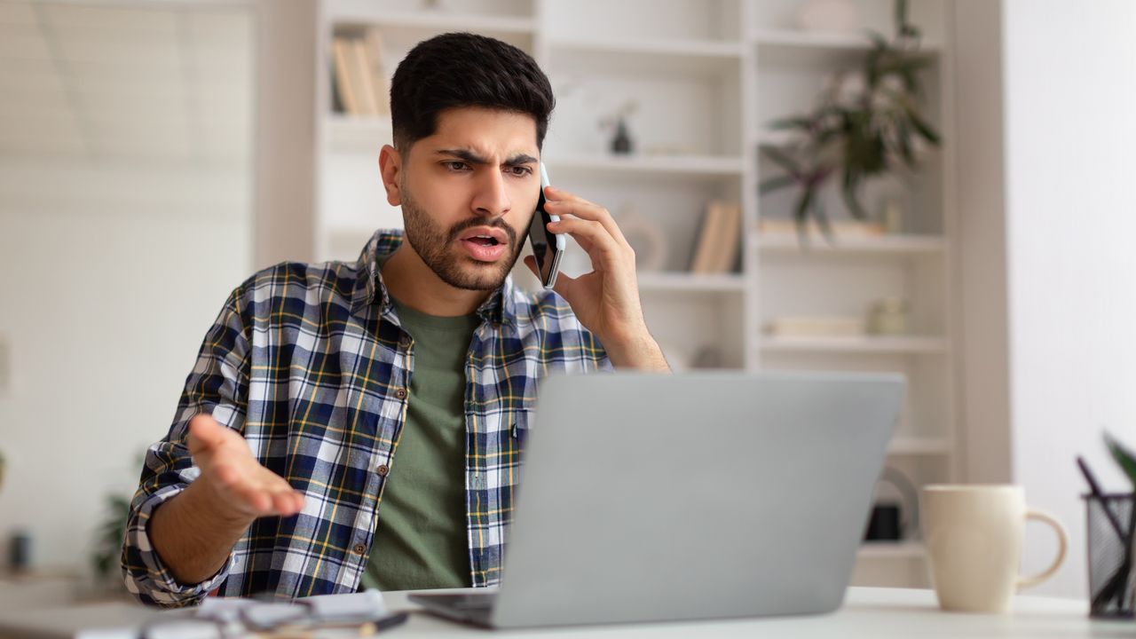 A young man looks annoyed as he talks on the phone while looking at his laptop.