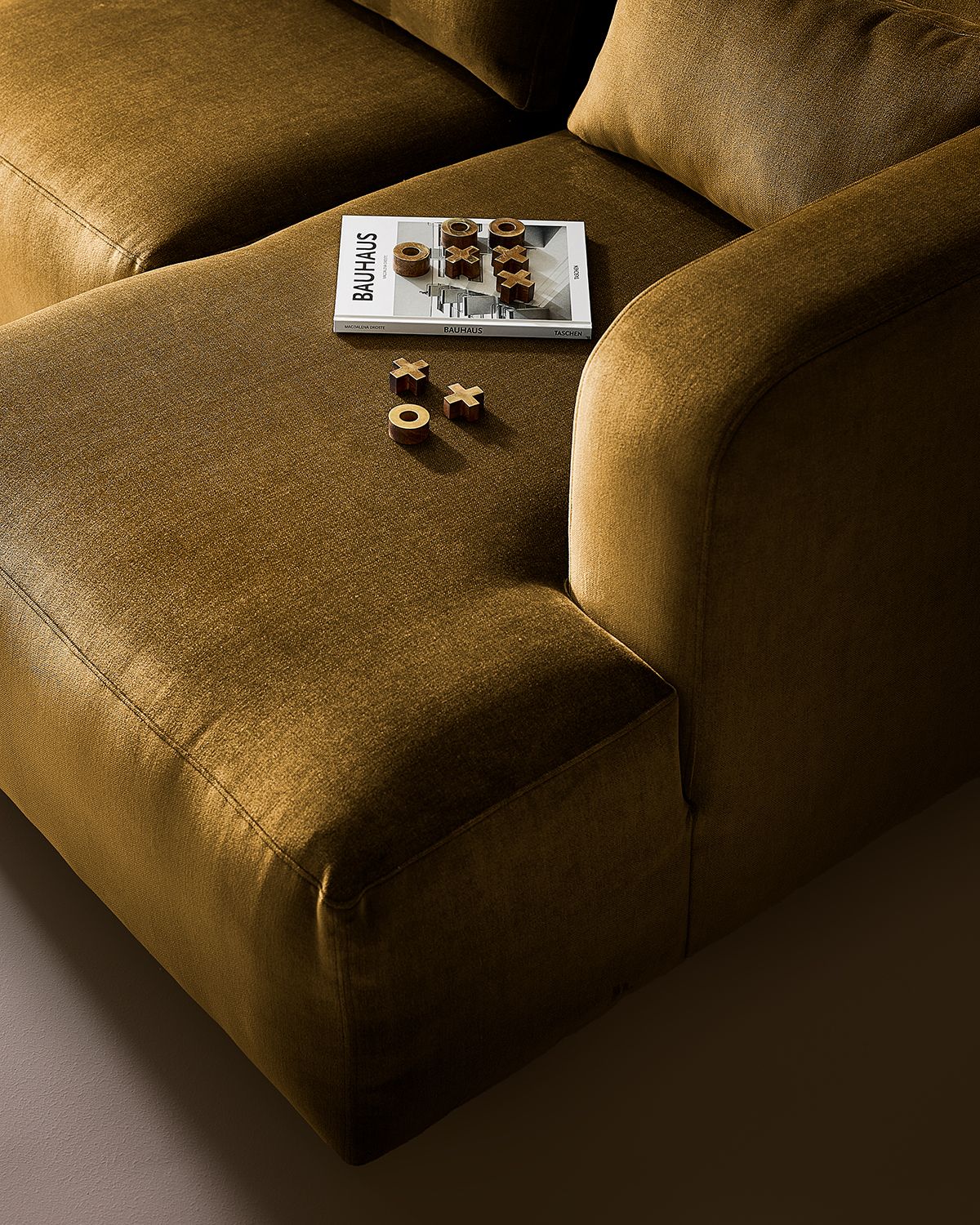 Close-up image of the lounge seat on a velvet, gold sofa. There is a book and a tic-tac-toe set of the seat as well.