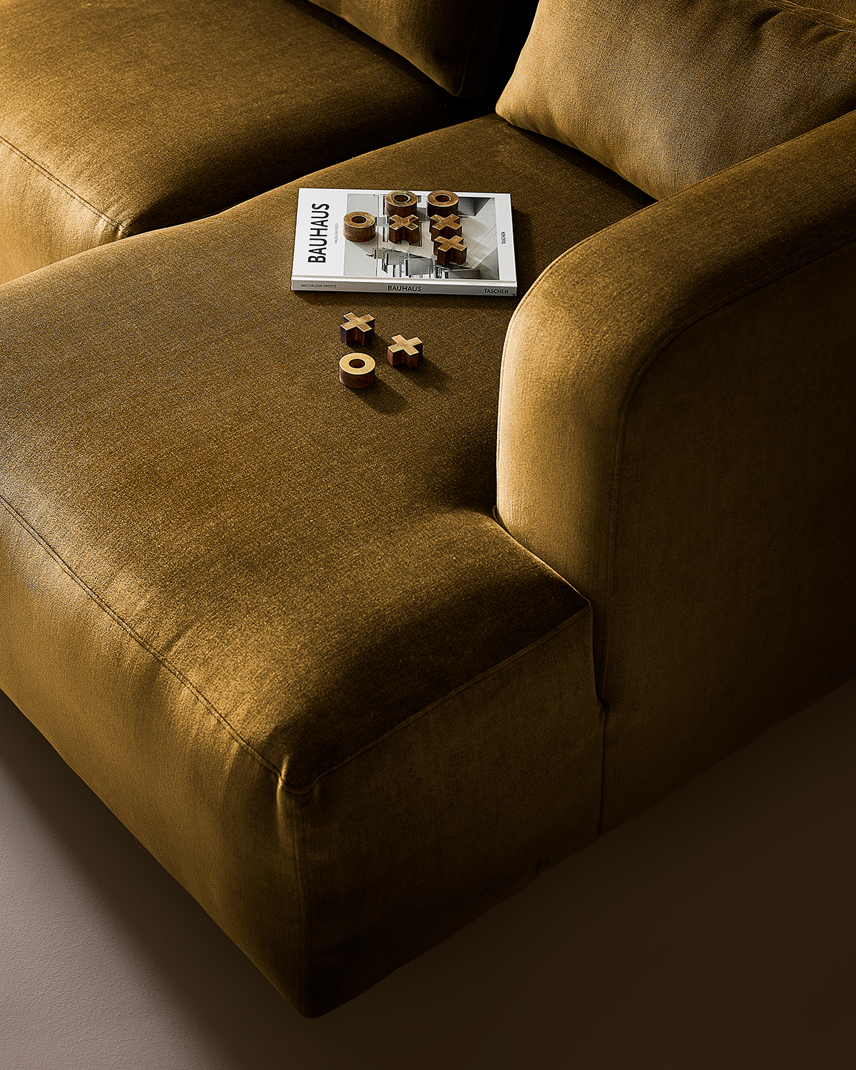 Close-up image of the lounge seat on a velvet, gold sofa. There is a book and a tic-tac-toe set of the seat as well.
