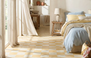 A sun-filled bedroom with a bold yellow and cream checkerboard tile floor flowing through to an open-plan ensuite. The bed is dressed in soft blue and yellow linen, beside a rustic wooden stool nightstand with a ceramic lamp. 