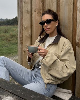 British influencer Jessica Skye sits at a cafe in Cornwall wearing black cay eye sunglasses, chunky hoop earrings, a cream cable knit sweater, tan barn coat, Khaite studded belt, and straight-leg jeans.