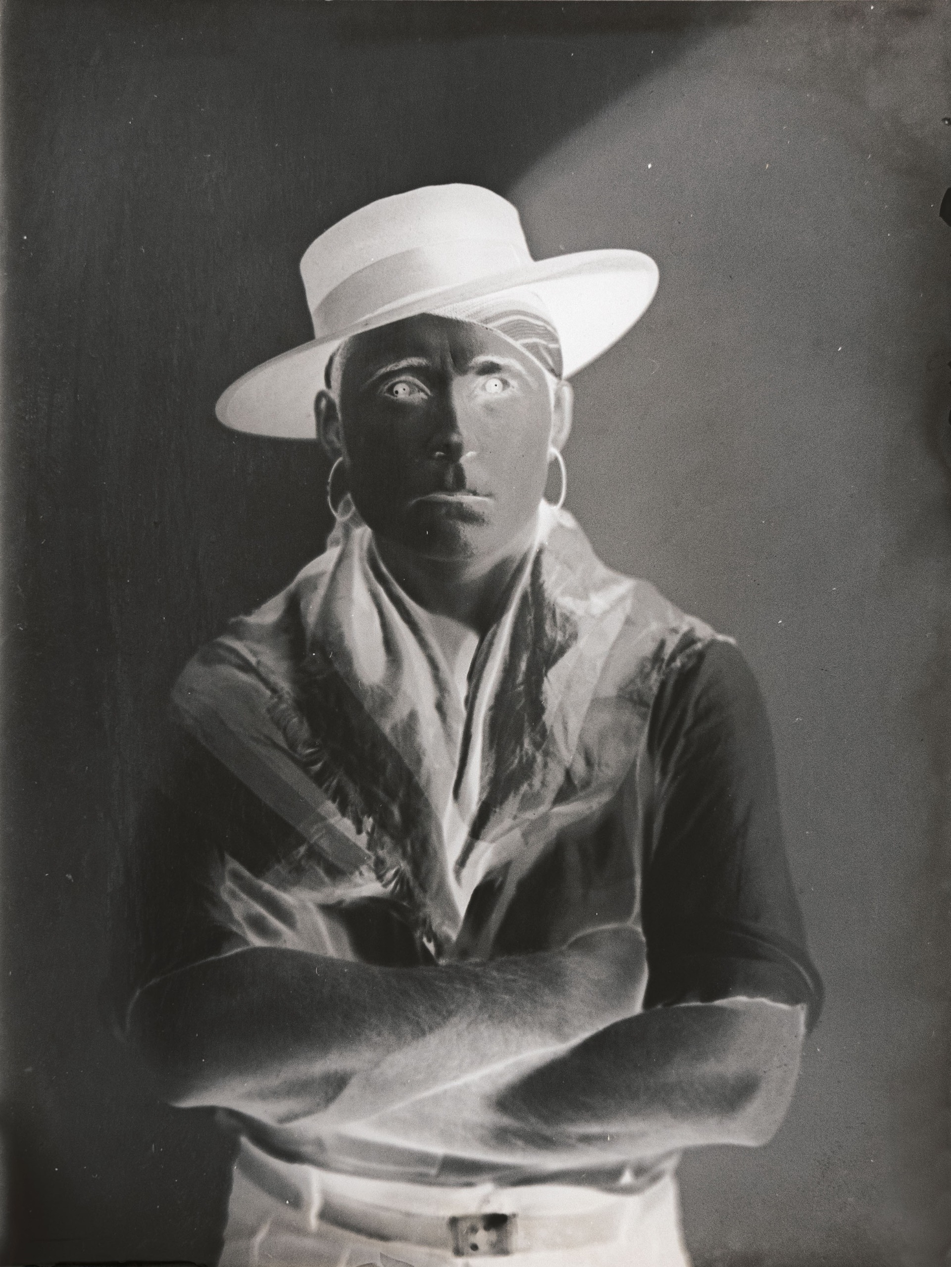 A double-exposed portrait of a man in a white boater hat and draped scarf, his face rendered dark with glowing white eyes.
