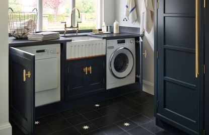 How to organize a laundry room, with black cabinetry and countertops, gold handles and faucet and a butler sink in between a washing machine and tumble dryer under a window.