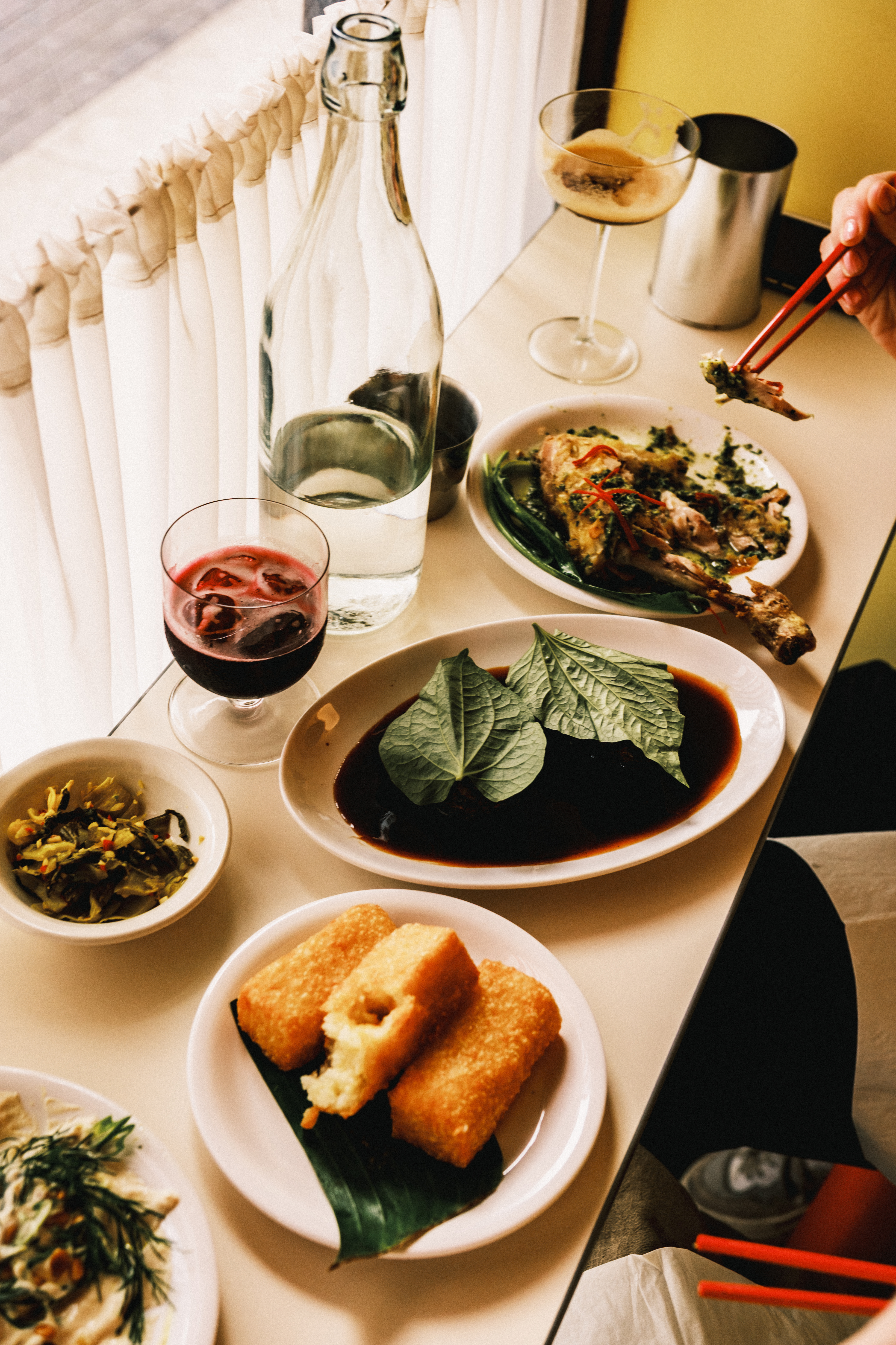A series of Asian food plates sits on a bistro-style wooden counter under cafe curtains along with a Martini and a glass of wine.
