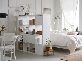 a white bedroom with a storage unit in the middle separating the bed from the white dinini g table